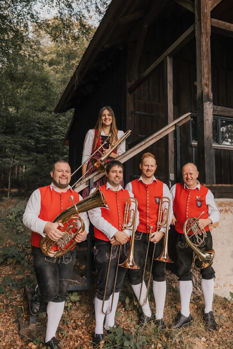Vier Männer in traditioneller Tracht mit roten Westen, weißen Hemden, schwarzen Kniebundhosen und weißen Strümpfen halten Blasinstrumente vor einer Holzhütte. Eine Frau in ähnlicher Tracht steht dahinter.