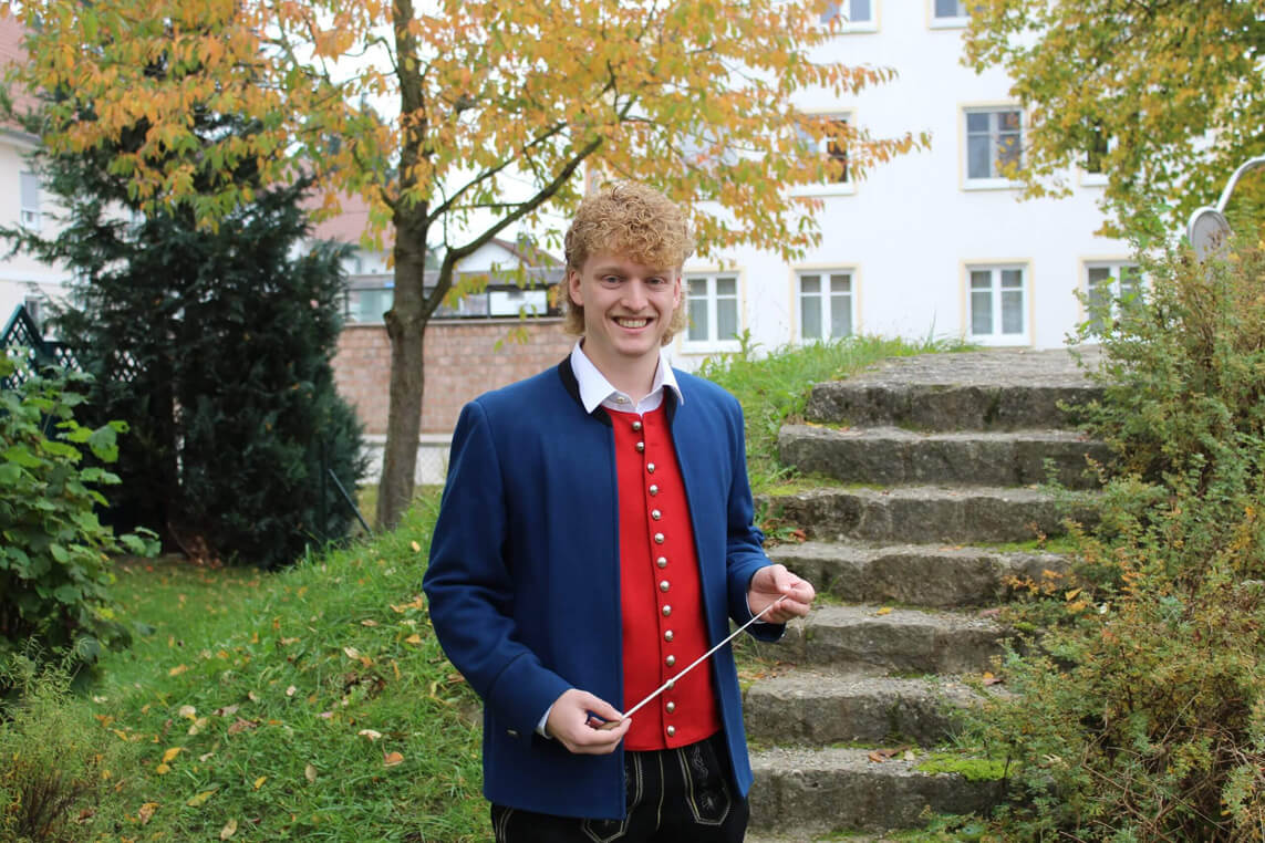 Christoph Mayer, Dirigent in traditioneller Trachtenjacke und rotem Pullover hält einen Dirigentenstab, steht vor einer Steintreppe im Garten.