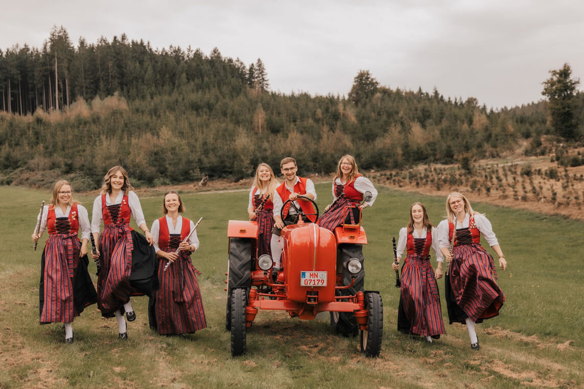 Mehrere Frauen in traditioneller Tracht laufen neben und hinter einem roten Traktor auf einer Wiese mit Wald im Hintergrund.