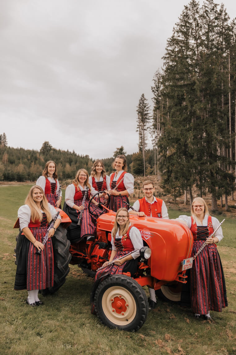 Gruppe von Musikerinnen und ein Musiker in Trachten mit Flöten und Klarinetten um einen roten Traktor auf einer Wiese vor Wald.