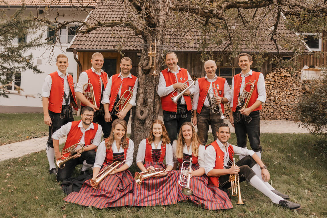 Gruppe von elf Personen in traditioneller Tracht mit roten Westen und karierten Röcken oder schwarzen Hosen, die Blasinstrumente halten, vor einem Haus mit Holzstapel.