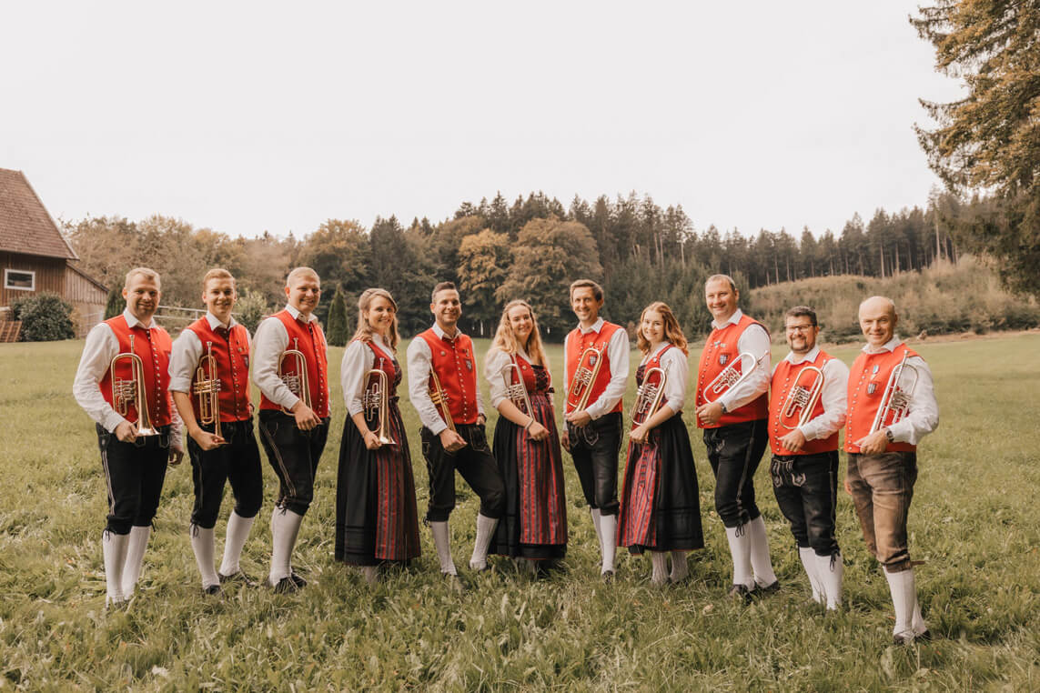 Gruppe von elf Personen in traditioneller Tracht mit roten Westen, weißen Hemden und schwarzen Lederhosen oder langen Röcken, einige halten Blasinstrumente, stehen auf einer Wiese vor Wald.