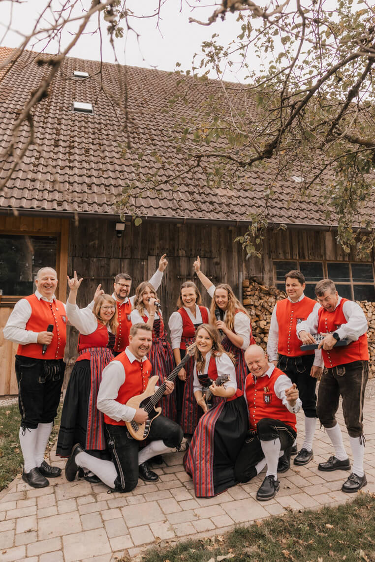 Gruppe von Menschen in traditioneller Tracht mit roten Westen und schwarzen Hosen oder Röcken, einige halten Musikinstrumente, vor einem Holzhaus mit Ziegeldach.