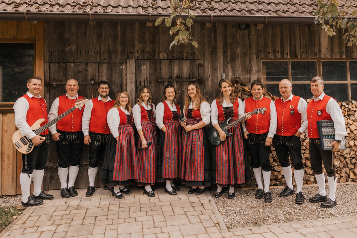 Gruppe von Menschen in traditioneller Tracht mit roten Westen, gestreiften Röcken und Musikinstrumenten vor Holzwand.
