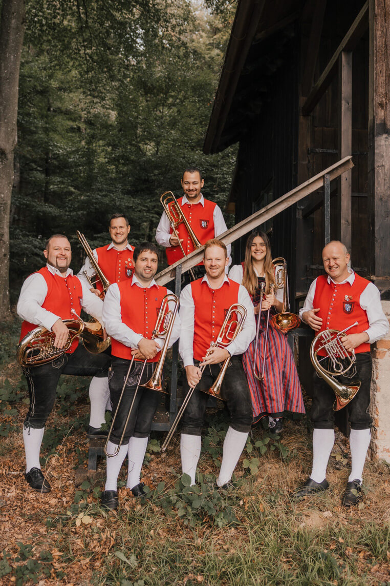 Sieben Personen in traditioneller Tracht mit roten Westen und weißen Hemden, halten Posaunen, zwei halten ein Horn, stehen vor einem Holzhaus im Wald.