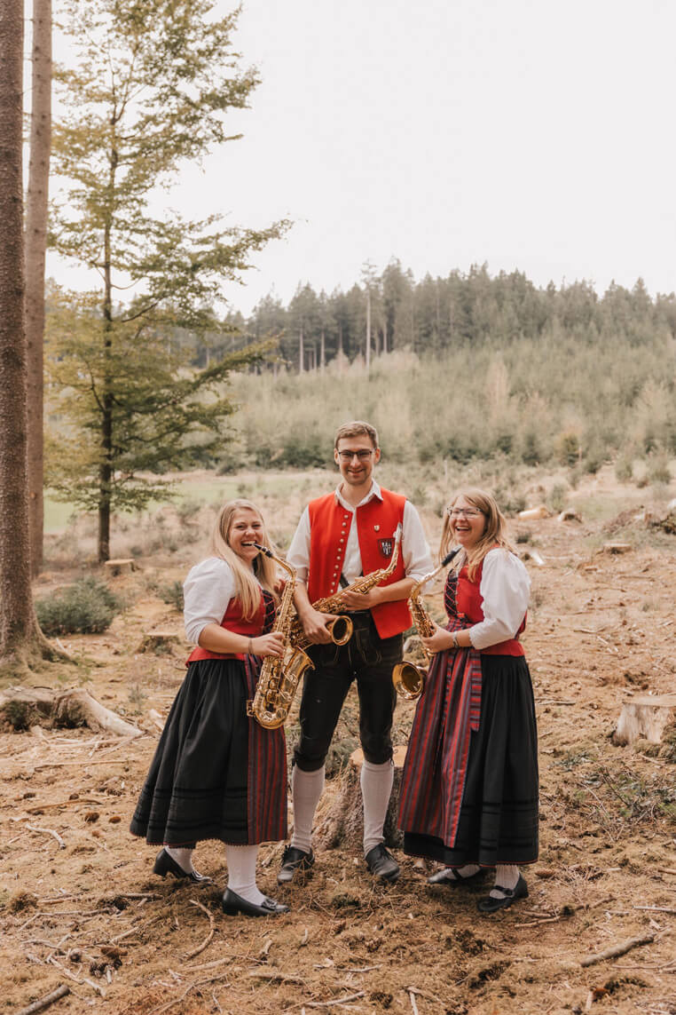 Drei Personen in traditioneller Tracht stehen im Wald und spielen Saxophone.