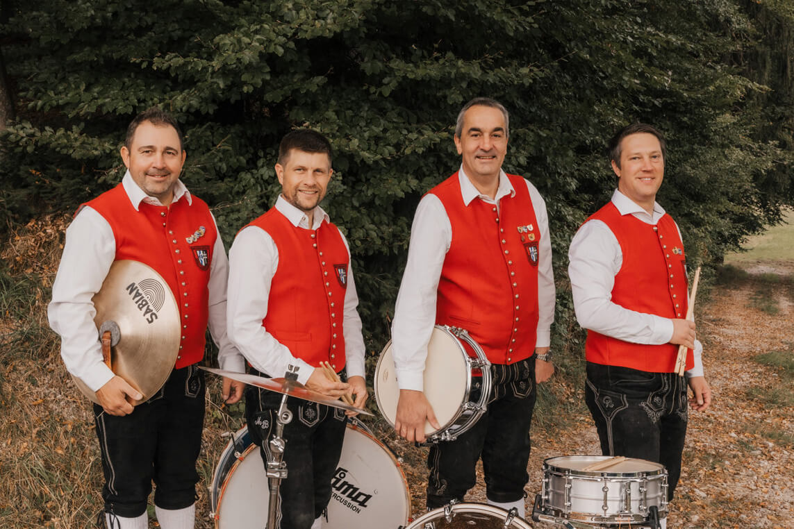 Vier Männer in roten Trachtenwesten und weißen Hemden stehen mit Schlaginstrumenten vor Büschen im Freien.