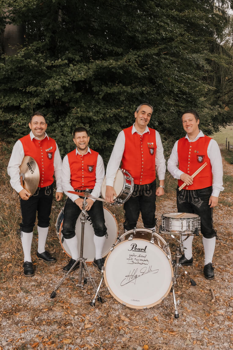 Vier Männer in roten Trachtenwesten, weißen Hemden und schwarzen Lederhosen stehen mit Schlaginstrumenten vor einem Baum.