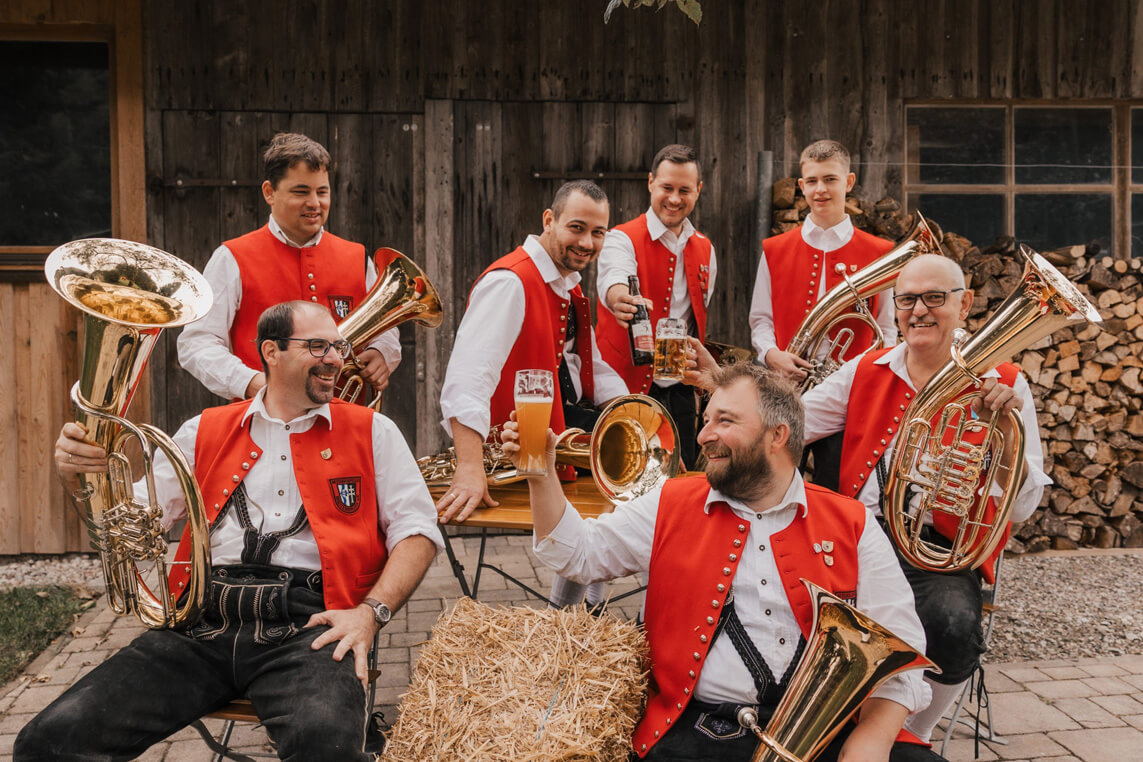 Sieben Männer in roten Trachtenwesten sitzen und stehen mit Tenorhörnern und Baritons vor einer Holzwand und halten Biergläser, Heuballen und Holzstapel im Hintergrund.