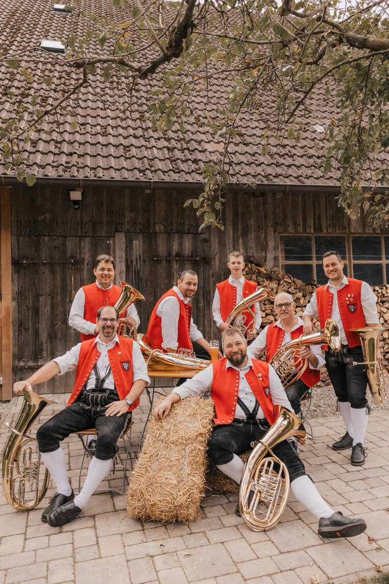Sieben Männer in traditioneller Tracht mit roten Westen und schwarzen Kniebundhosen posieren mit Tenorhörnern und Baritons vor einem Holzgebäude und einem Heuballen.