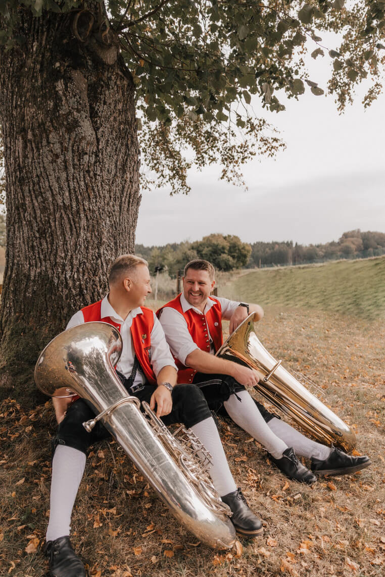 Zwei Männer in traditioneller Tracht mit roten Westen sitzen unter einem Baum und halten je eine Tuba.